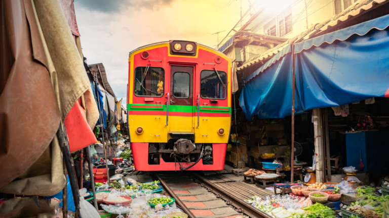A bright red and yellow train sits on tracks in a crowded market street, flanked by colorful stalls selling vegetables and goods on both sides.