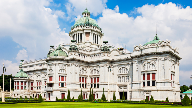 Grand white palatial building with green copper domes and a manicured foreground garden against a bright blue sky.