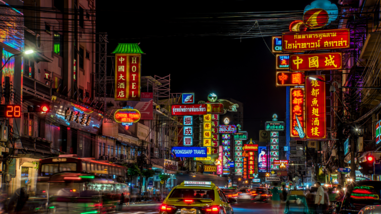 Bustling Chinatown street at night with bright neon signs in Chinese and Thai, traffic including taxis and blurred vehicles.