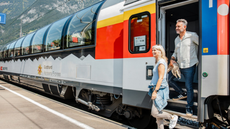 Two passengers step off a red-yellow Gotthard Panorama Express train onto a platform with mountains in the background, smiling as they exit.