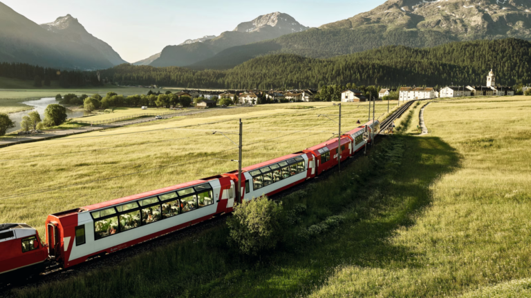 Red passenger train winds through a golden valley with a small village, trees, and distant mountains.