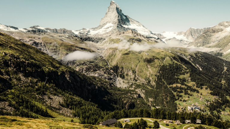 Snowy alpine peak rises above green valleys and a small village at the base under a clear blue sky.