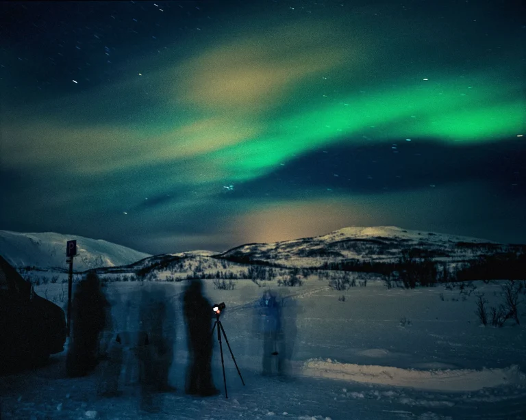 Aurora borealis glows green across a dark night sky above a snow-covered landscape with silhouettes of people and a camera on a tripod in the foreground.