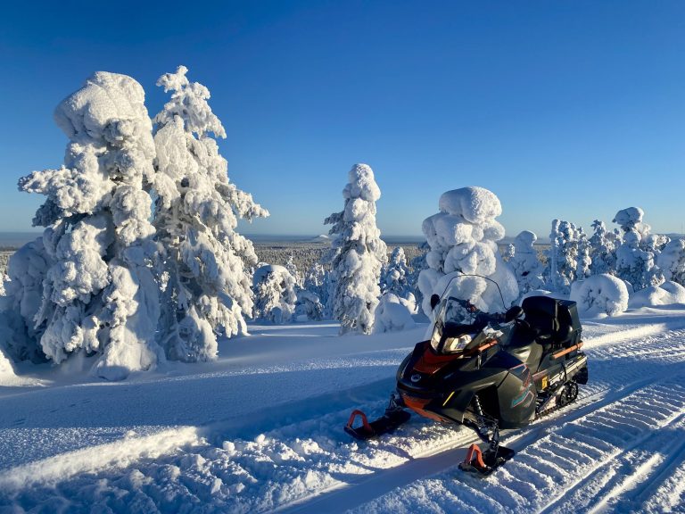 Snow-covered trees under a blue sky with a snowmobile in the foreground.