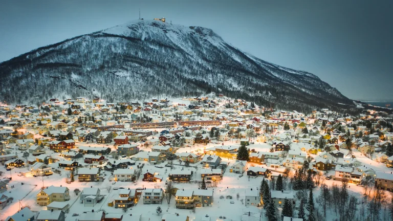 Snow-covered mountain rises above a small town with warmly lit houses at dusk/dawn, lights dotting the snowy landscape.