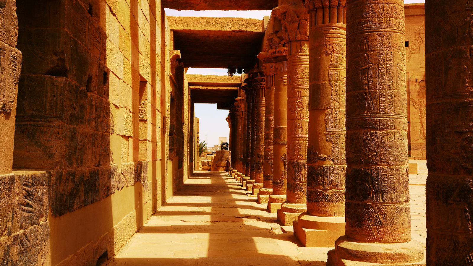 Sunlit ancient Egyptian colonnaded corridor with carved hieroglyphs on sandstone pillars.