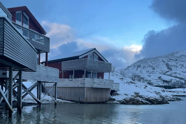 Red wooden cabins on stilts along a calm, snow-covered shoreline with blue sky and distant mountains in view.