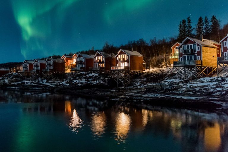 Row of red wooden cabins on stilts along a snowy shoreline under the green glow of the aurora borealis.