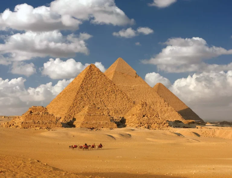 Three ancient Egyptian pyramids rise from a sandy desert under a partly cloudy blue sky; a line of camels travels in the foreground.