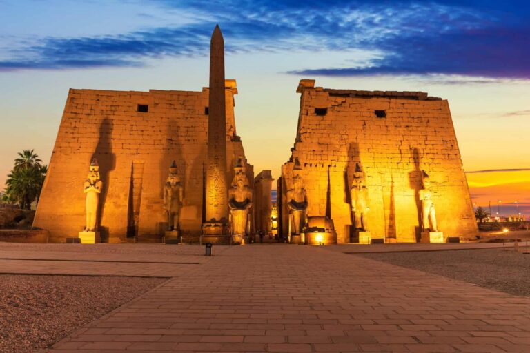 Egyptian temple complex at sunset with a central obelisk and statues lit by golden lights