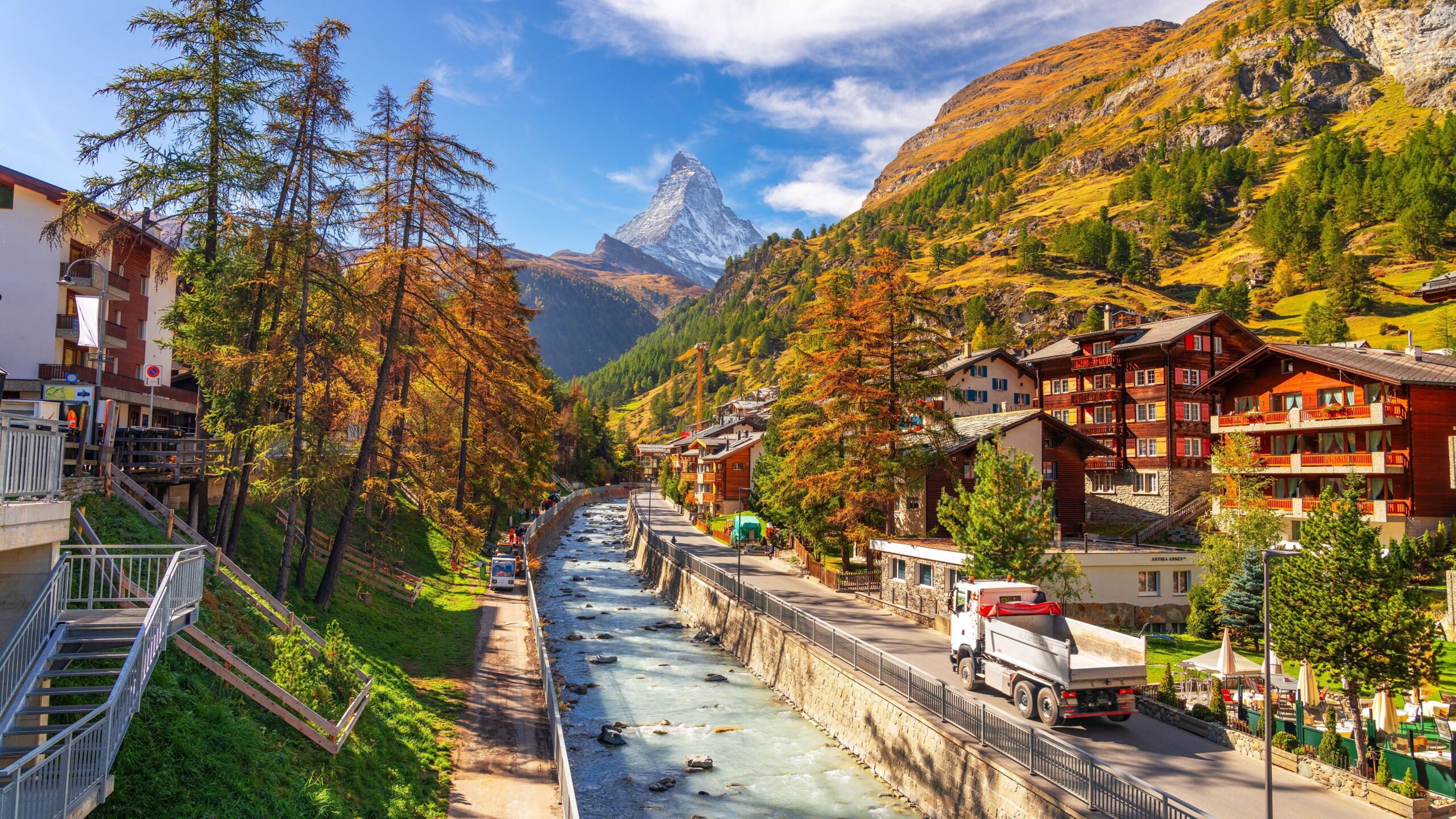 Alpine village with wooden chalets along a river valley, trees changing color, and a snow-capped peak in the background.