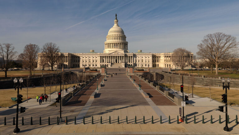 Front view of the U.S. Capitol with its dome, flanked by wings, seen from a wide plaza and walkway on a clear day, leafless trees nearby.