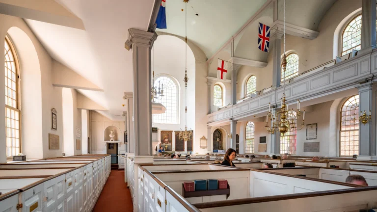 Interior of a historic church with white pews, arched windows, and ornate brass chandeliers; flags hang from the balcony above. UX: worship space in daylight.