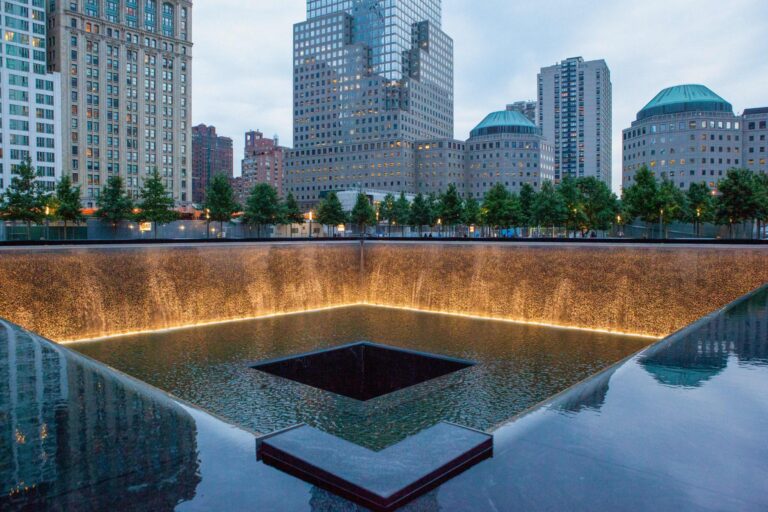 Evening view of the 9/11 Memorial reflecting pool with cascading gold-lit water and a city skyline in the background.