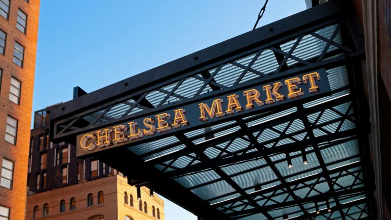 Chelsea Market entrance sign hanging from a black metal framework against a blue sky.