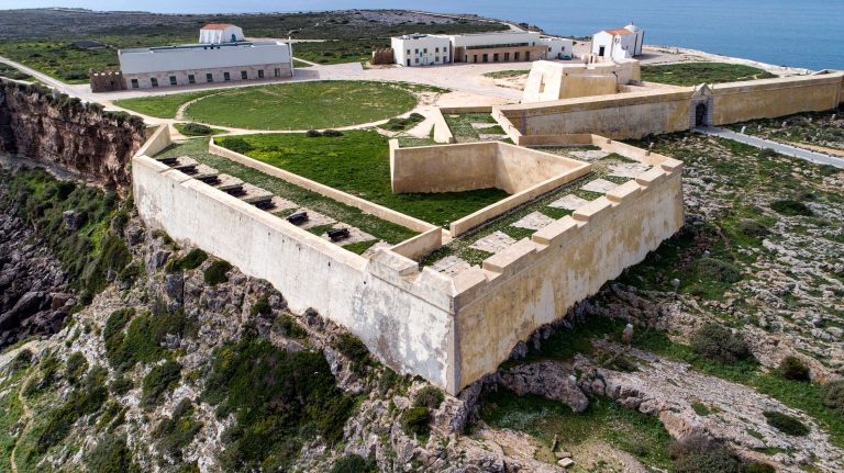 Aerial view of a coastal fortress with beige stone walls, cannons along the ramparts, and buildings by the sea.