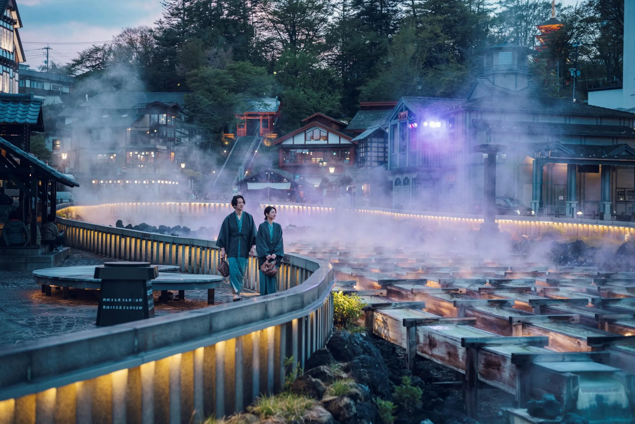 Two people in traditional blue robes walk along a lit boardwalk beside a steaming hot spring in a Japanese onsen town.