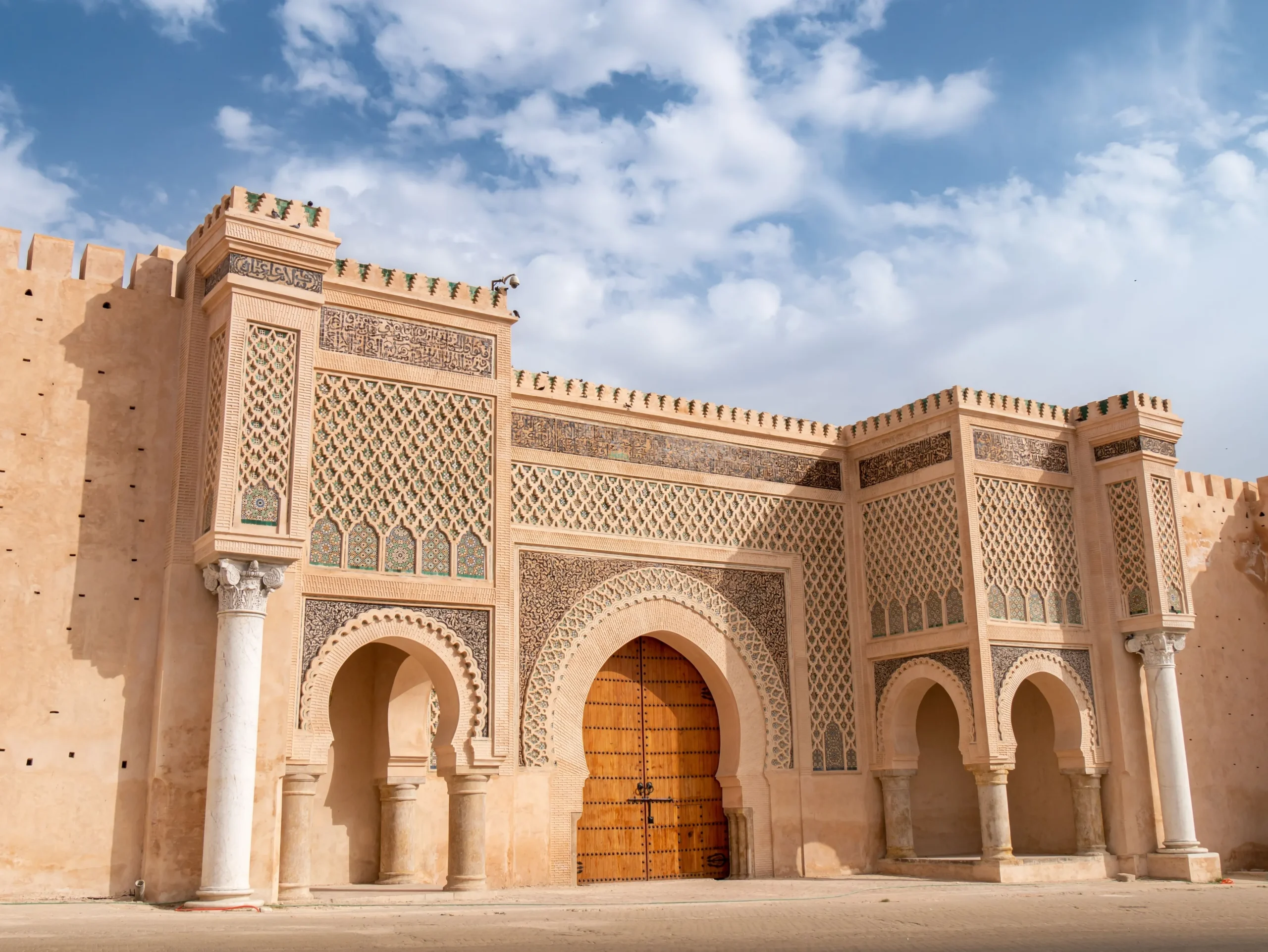 Ornate sandstone facade with geometric tilework, arches, and columns under a blue sky.