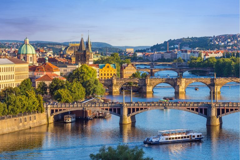 Panoramic view of Prague with the Charles Bridge over the Vltava River, historic buildings, and green hills in the distance, daytime light.