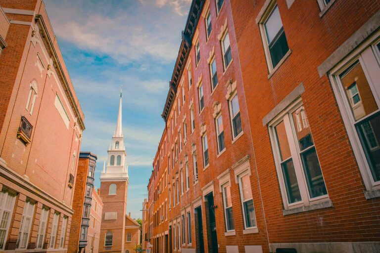 Narrow brick alley flanked by red brick buildings, with a white church steeple at the end against a blue sky.