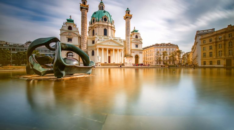 Baroque Karlskirche church with a green dome and tall columns, reflected in a calm pool with a modern sculpture in the foreground