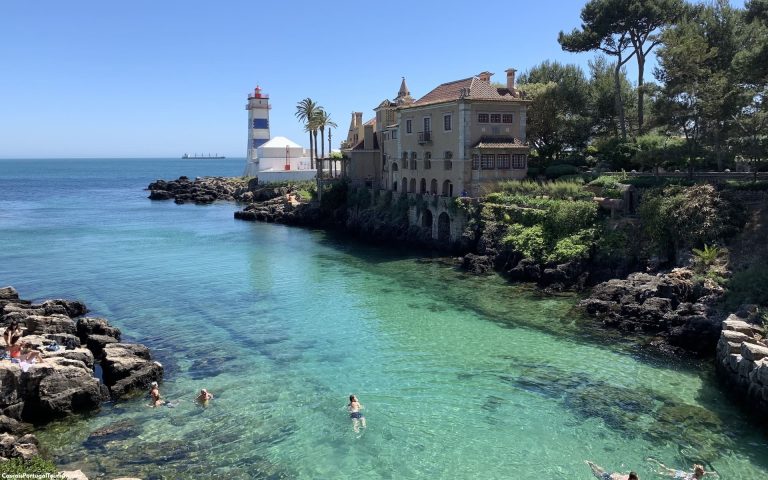 Turquoise water along a rocky shore with a white lighthouse and a historic villa on the coast under a blue sky.