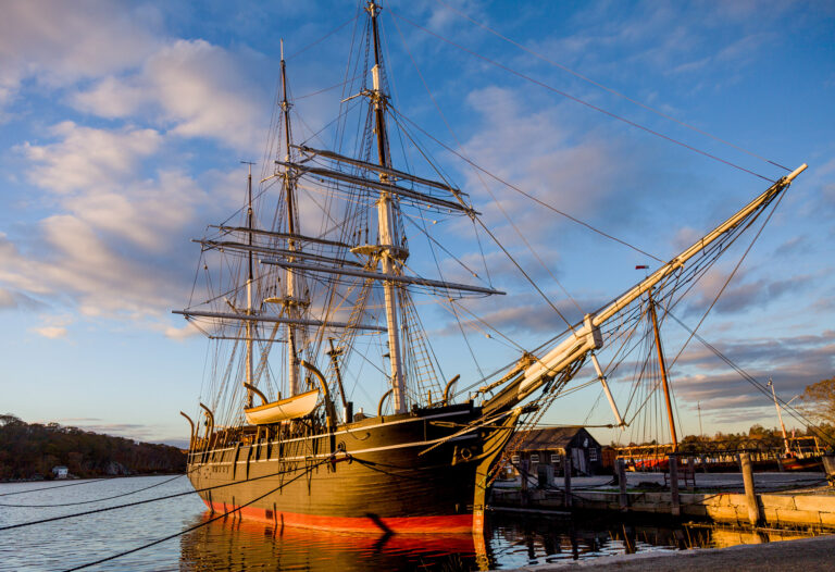 Tall ship with multiple masts and rigging docked at a marina at sunset.