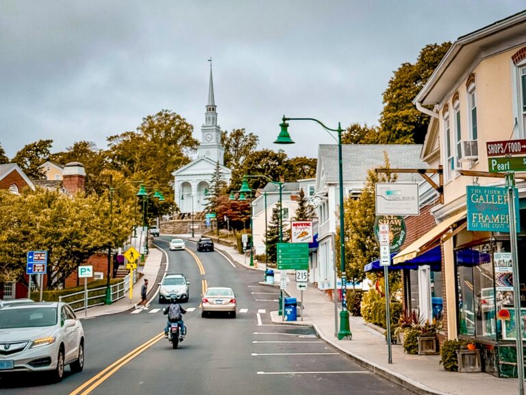 Street view of a small town with a white church steeple, green lampposts, and storefronts on the right under a cloudy sky.