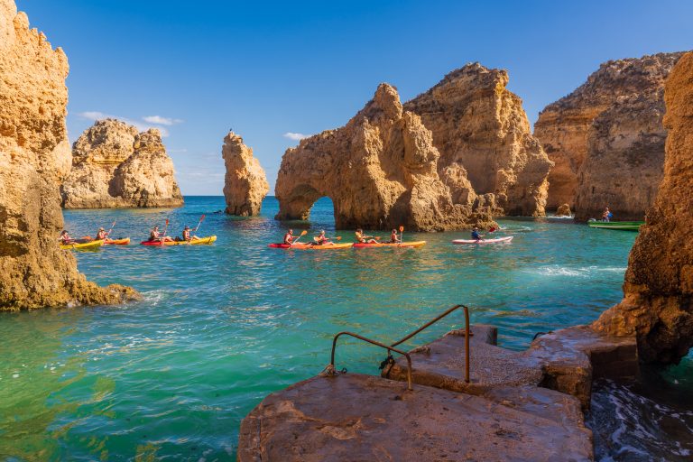 Group of kayakers paddling in turquoise water between towering orange rock arches under a clear blue sky.