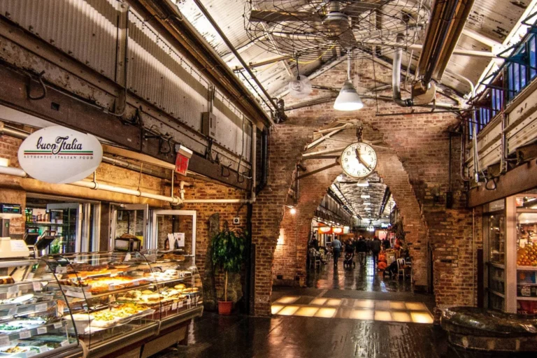 Indoor market corridor with brick arches, a deli counter on the left, and a hanging clock under an industrial ceiling.