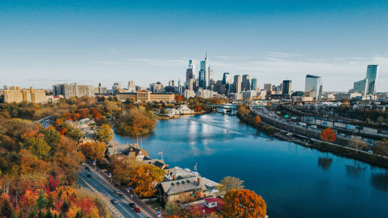 City skyline along a calm river with autumn trees in the foreground and a clear blue sky above.