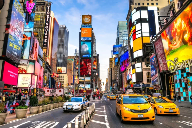 Busy Times Square street with yellow taxis, pedestrians, and colorful illuminated billboards everywhere.",