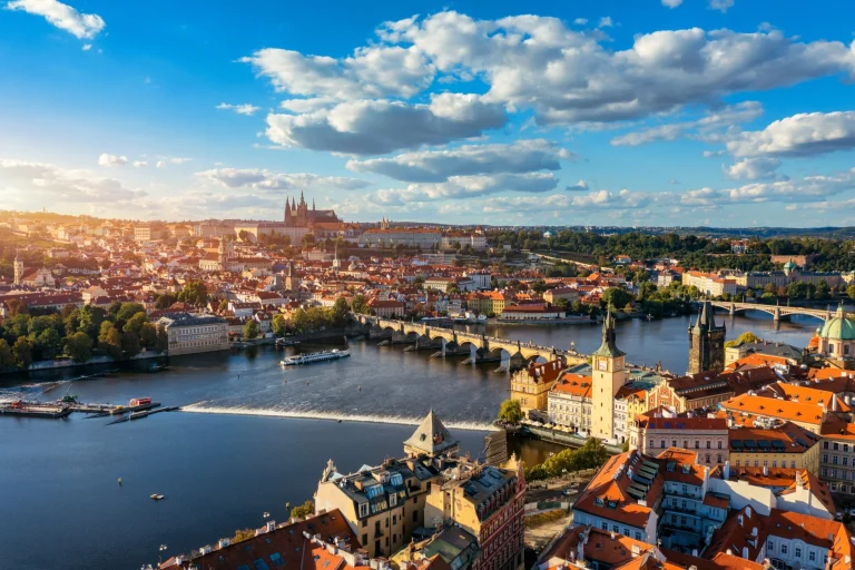 Aerial panorama of Prague with red-roofed buildings, a river, and a stone bridge under a blue sky with fluffy clouds.