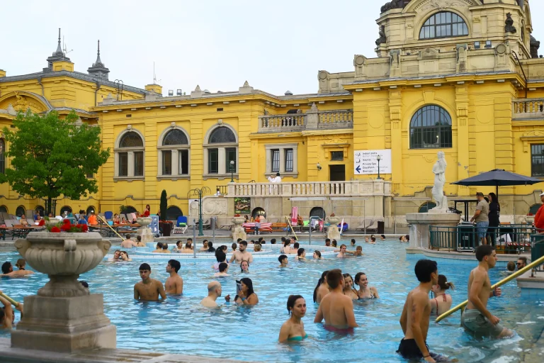 Outdoor thermal pool crowded with swimmers in front of a bright yellow historic building with arched windows and a statue plaza.