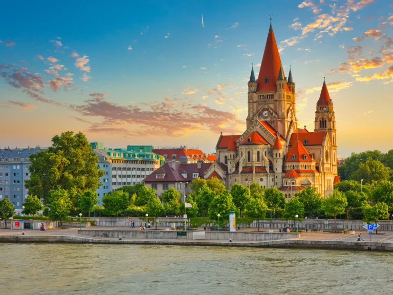 Riverside cityscape at sunset with a large Gothic church featuring bright red spires on the right.